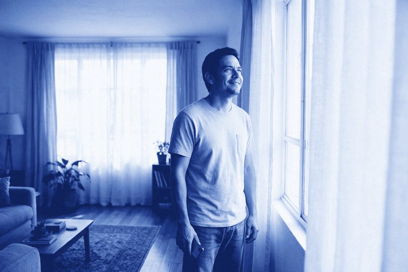 Hombre adulto de pie junto a la ventana de su sala, sereno, con el teléfono en la mano.